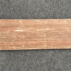 Foto einer einzelnen Bodenfliese Feinsteinzeug im Format 22,5cm x 90cm. Diese Fliese hat einen rötlichen, anpolierten Braunton gehalten, der teilweise glänzend und teilweise matt erscheint. Die Fliese hat eine Holzoptik, die kaum von echtem Holz zu unterscheiden ist.